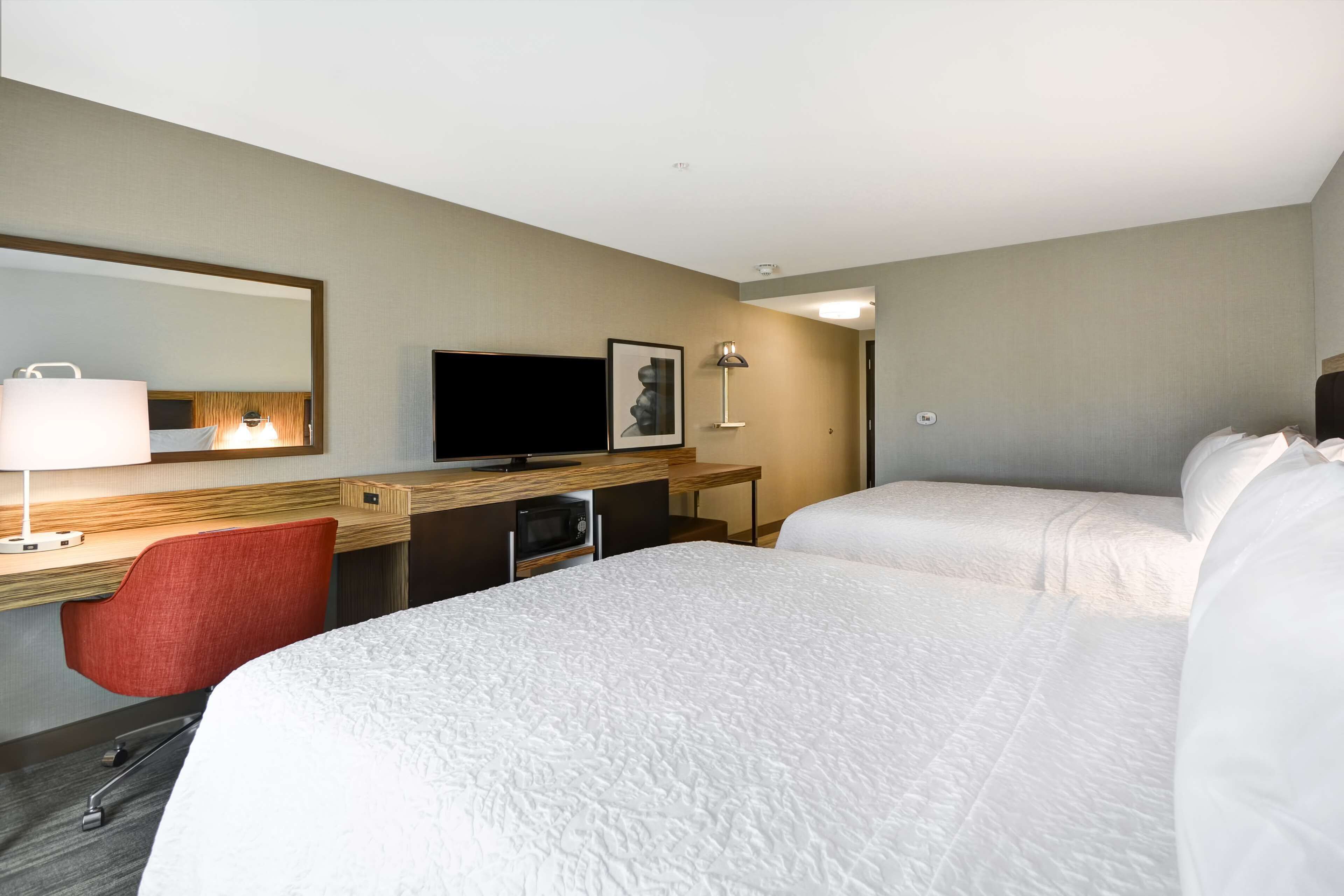 Ett hotellrum med två sängar, ett skrivbord med en stol, en TV, en lampa och en spegel.