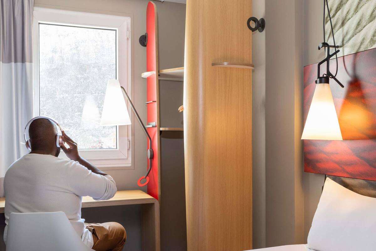 A hotel room with a bed, a desk, a window, and a red surfboard.