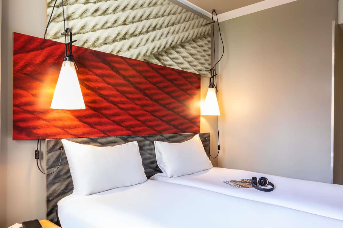 A hotel room with a bed, two bedside lamps, a headboard with a textured design, a nightstand with a phone and book, and a wall with a large abstract painting.