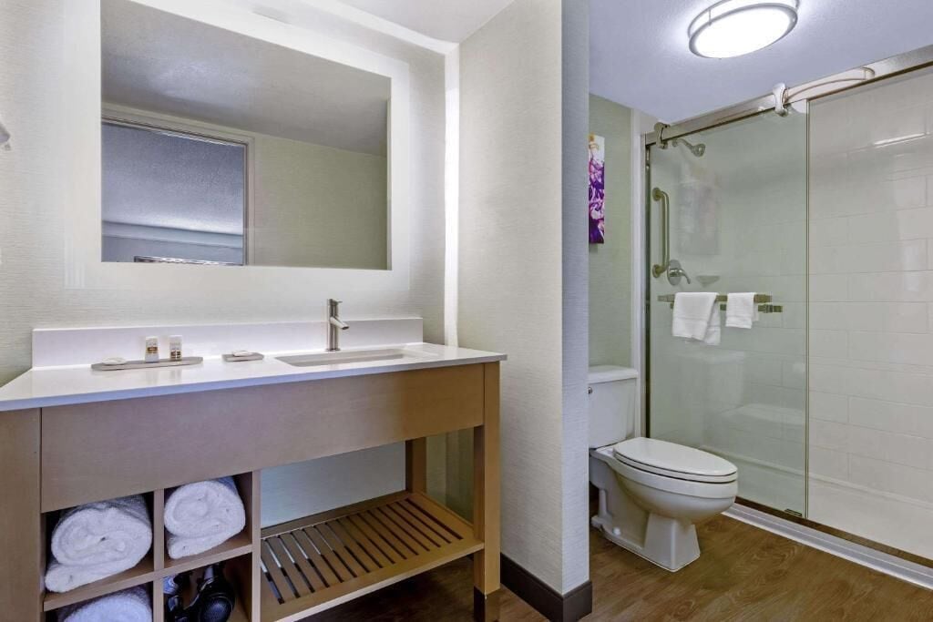 Une salle de bain moderne avec une douche à parois de verre, un lavabo blanc avec un miroir au-dessus et un meuble-lavabo en bois agrémenté de serviettes.