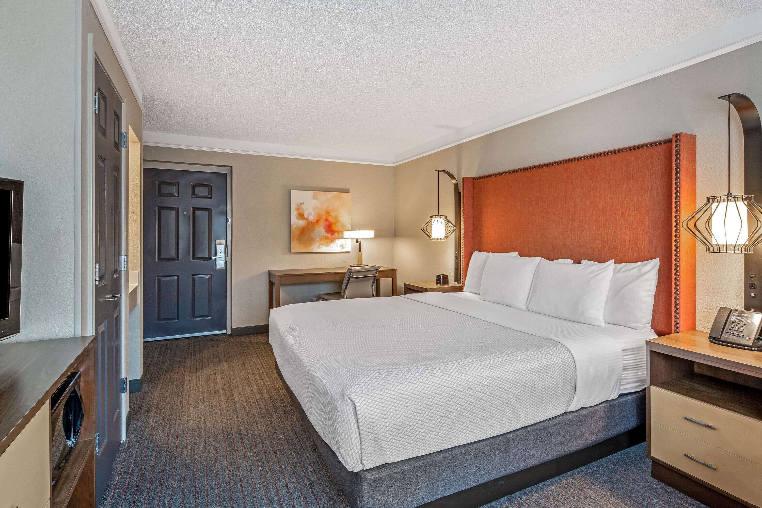 A hotel room with a large bed, a desk with a chair, a desk lamp, a telephone, and a television.