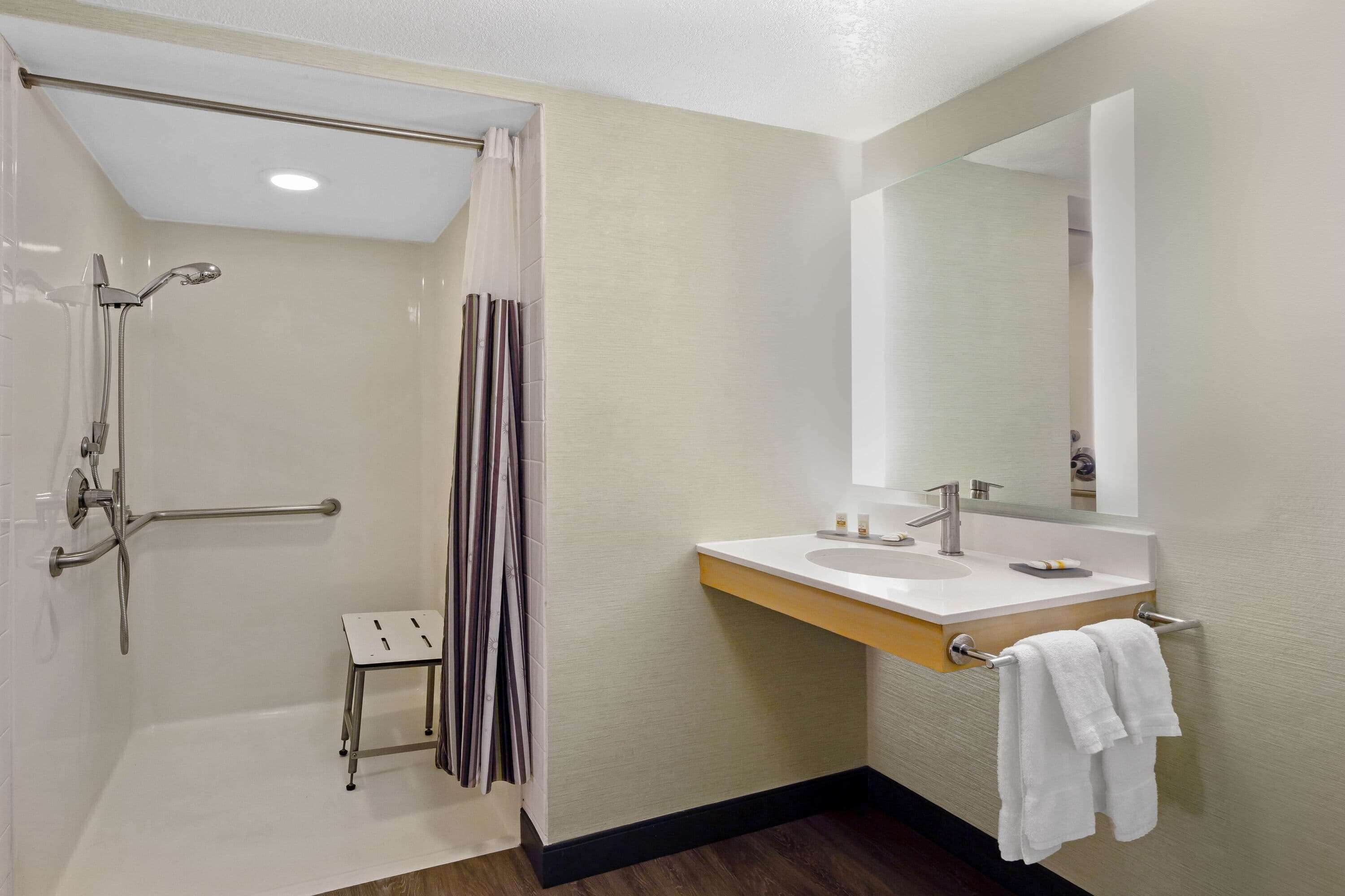 Un baño moderno con ducha, un taburete pequeño, tocador con lavamanos y espejo.