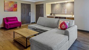 Un salon moderne avec un canapé gris, une table basse en bois et un fauteuil rose vif. On aperçoit à l’arrière-plan un coin cuisine avec un plan de travail et un évier.