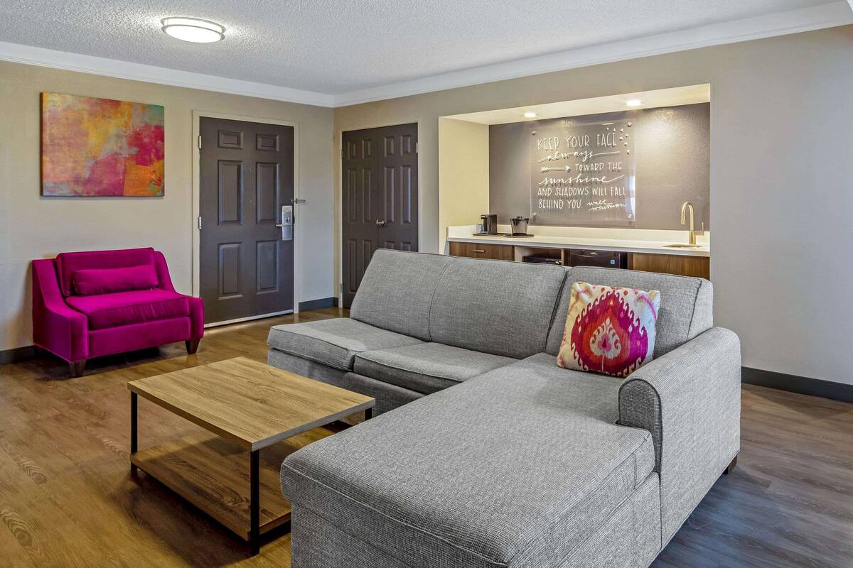 Una sala moderna con un sofá gris, una mesa de centro de madera y un sillón rosa vibrante. Se observa un área de cocina con una encimera y un fregadero al fondo.