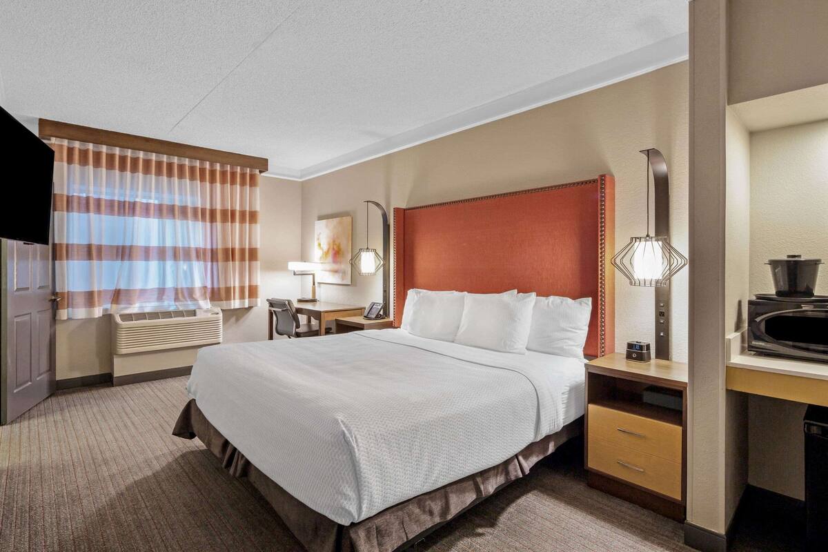 Una habitación de hotel con una cama grande, un escritorio con computadora, televisor y ventana con cortinas a rayas.