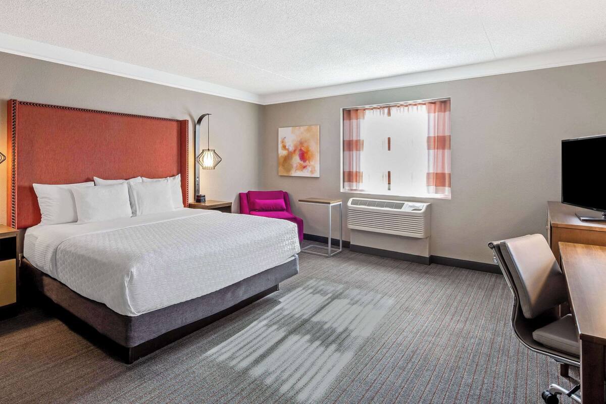 Habitación de hotel con una cama grande, un escritorio, una silla, un televisor, una ventana con cortinas y un radiador.
