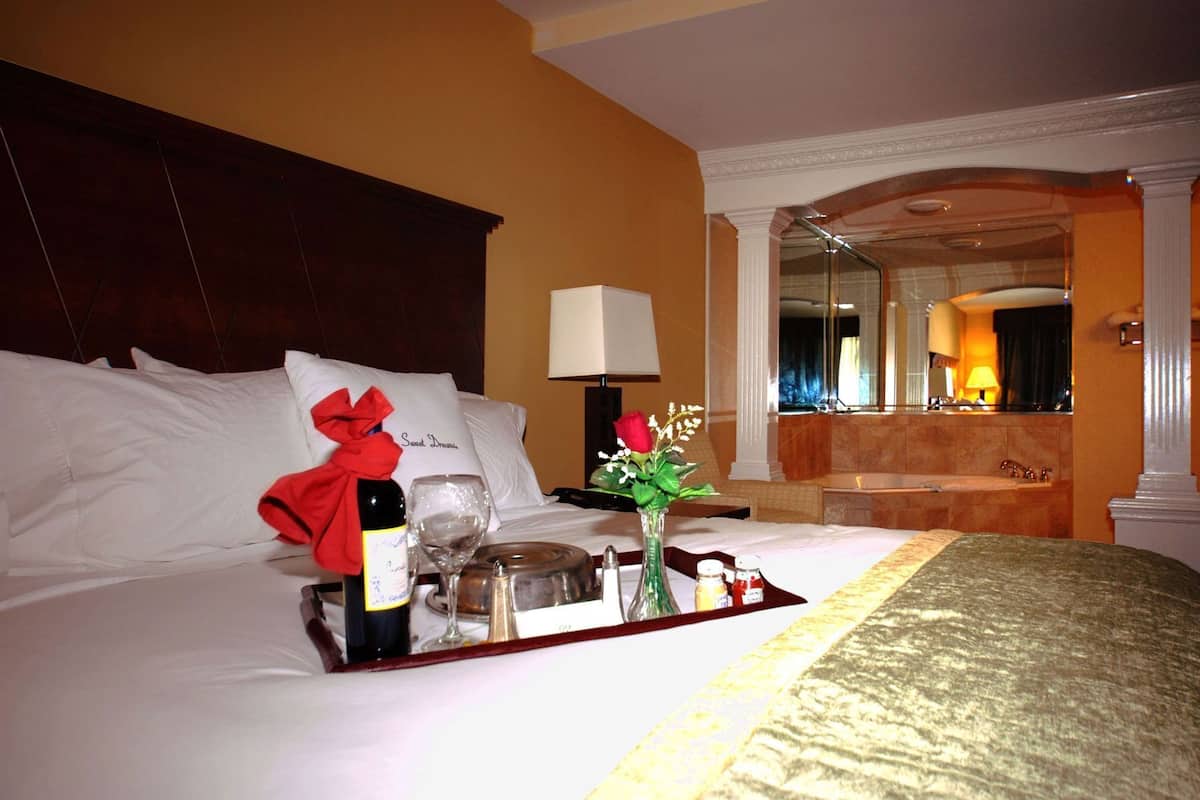 A hotel room with a neatly made bed, a tray with a bottle, glasses, and a vase with flowers.