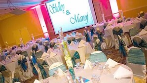 A banquet hall with round tables covered in white tablecloths, each set with silverware and glassware. The room is decorated with blue and white drapery and has a stage with a screen displaying "Oshkalla & Manji".