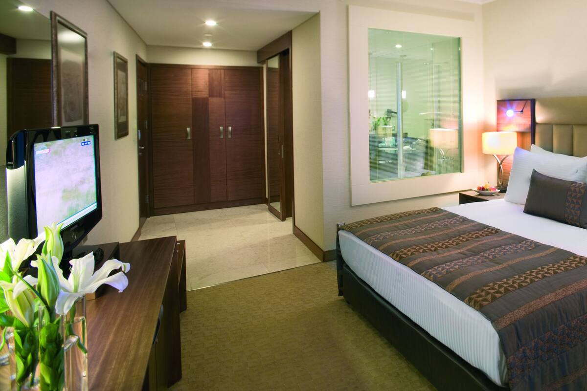 Habitación de hotel con cama, lámparas de noche, televisor, escritorio con lámpara y ventana con persianas.