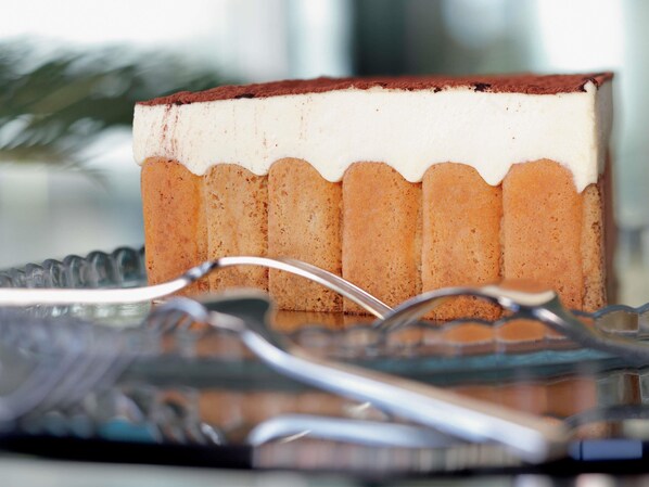 Una porción de pastelito en capas con relleno cremoso blanco y cobertura de chocolate, servido sobre un plato de vidrio transparente con una cuchara.