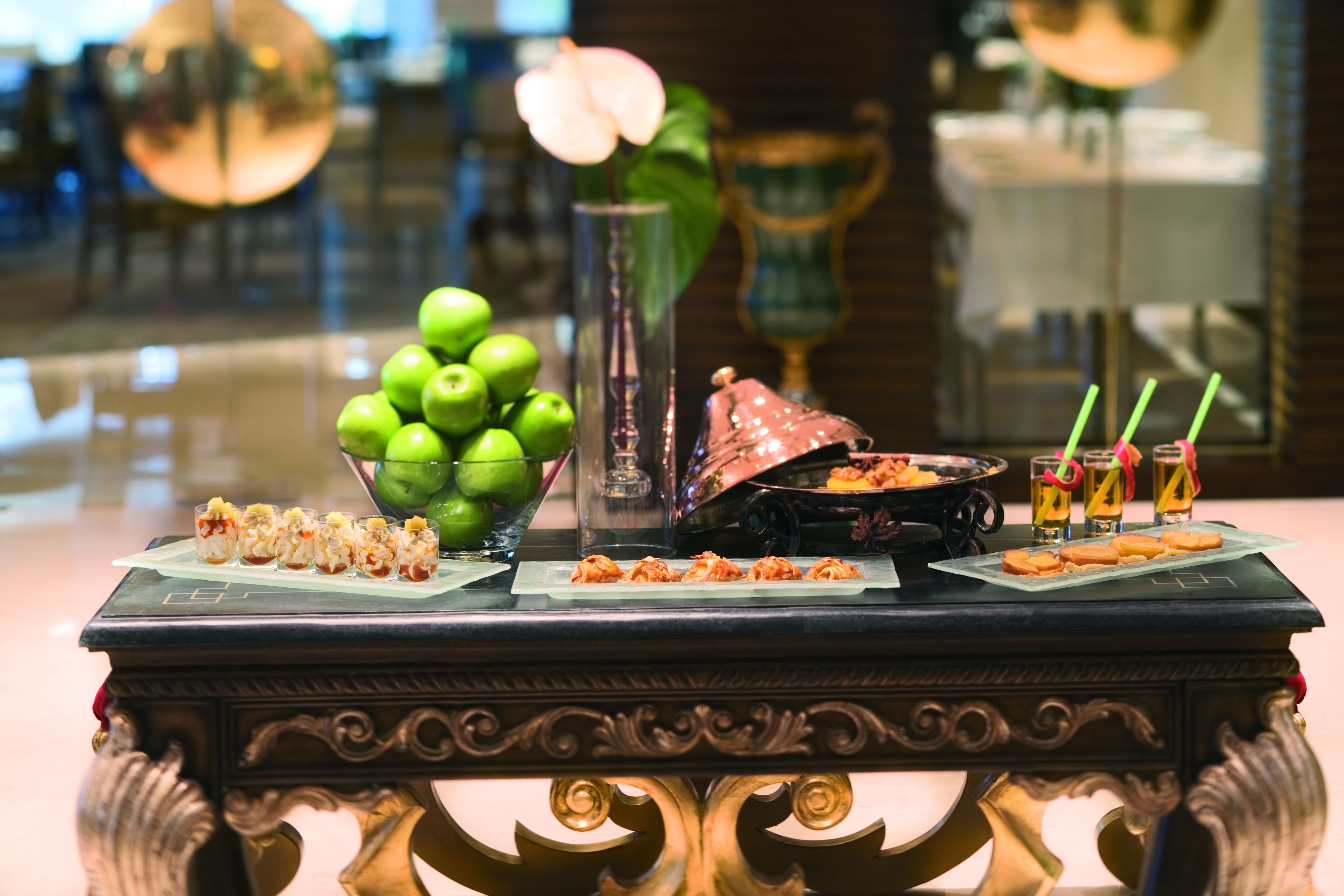 A table with various food items, including a bowl of green apples, a vase, and several plates of appetizers.