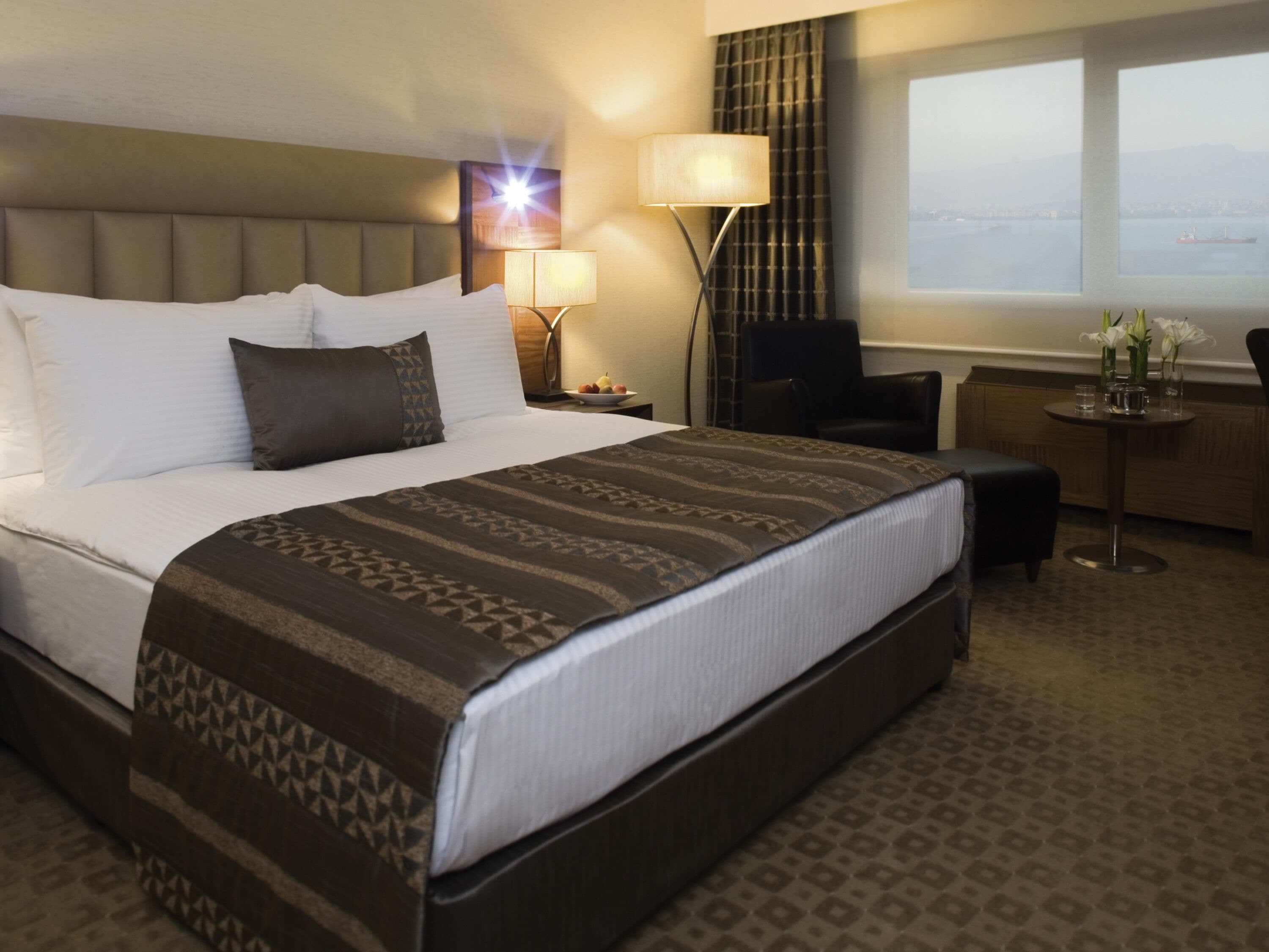 Ein Hotelzimmer mit einem großen Bett, einem Schreibtisch, einem Stuhl, einer Lampe und Meerblick.
