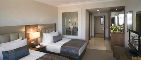 A hotel room with two beds, a desk, a TV, and a bathroom visible through a glass door.