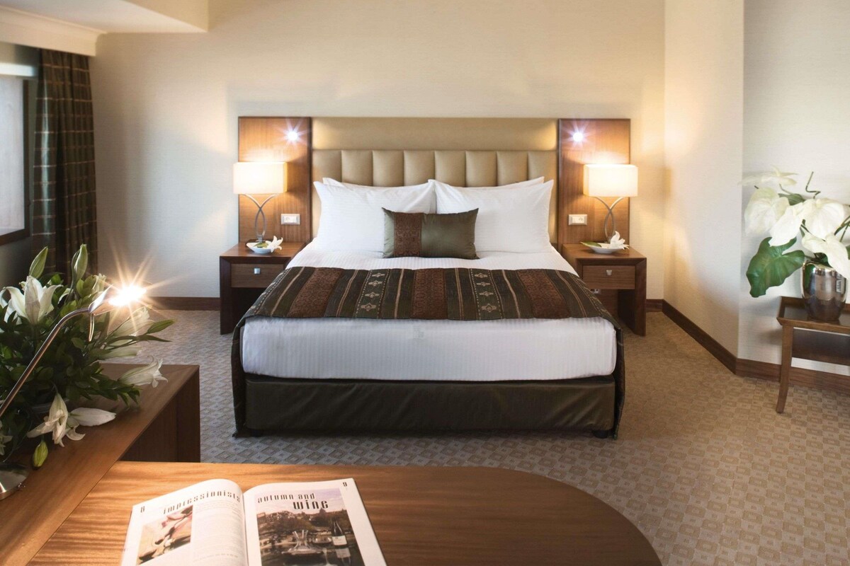 Habitación de hotel con una cama grande, dos mesitas de noche con lámparas, una mesita auxiliar con una lámpara, una planta en maceta y una revista sobre una mesita.
