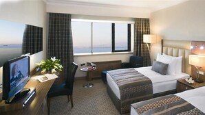 A hotel room with two beds, a desk with a TV, a small table with a chair, a window with a view, and a lamp. - Mövenpick Hotel Izmir (Izmir)