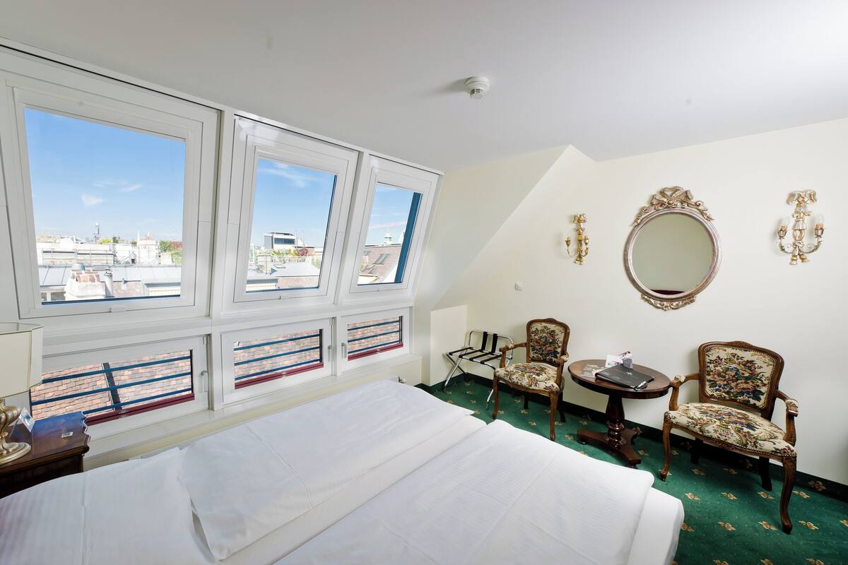 Hotellihuone, jossa on suuri sänky, kaksi tuolia, pieni pöytä, peili ja suuret ikkunat, joista avautuu näkymä kaupunkiin.