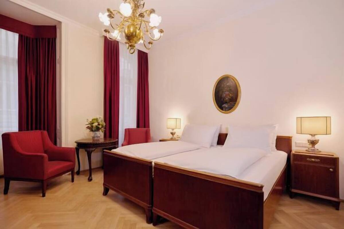 Hotellihuone, jossa on parivuode, kaksi yöpöytää valaisimineen, punainen nojatuoli, pieni pöytä maljakolla ja kattokruunu.