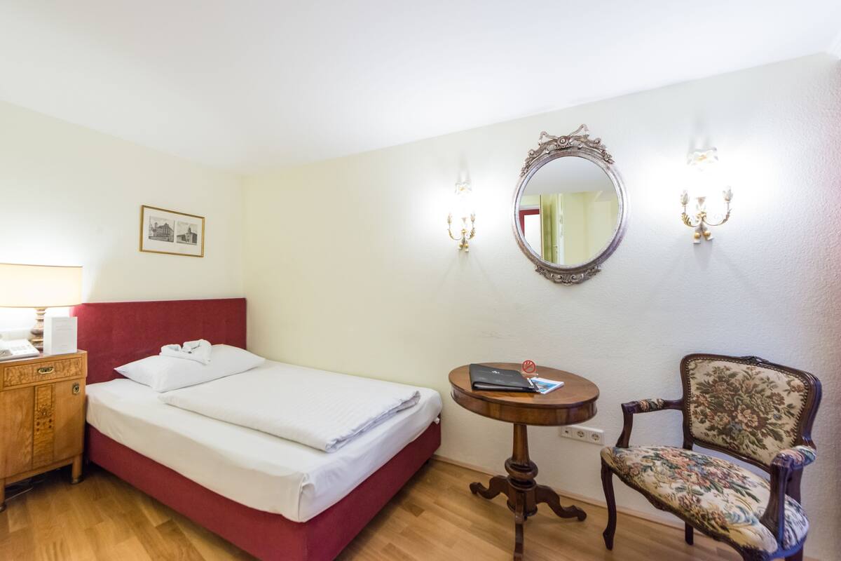 Hotellihuone, jossa on sänky, yöpöytä, tuoli, peili ja seinään kiinnitetty valaisin.
