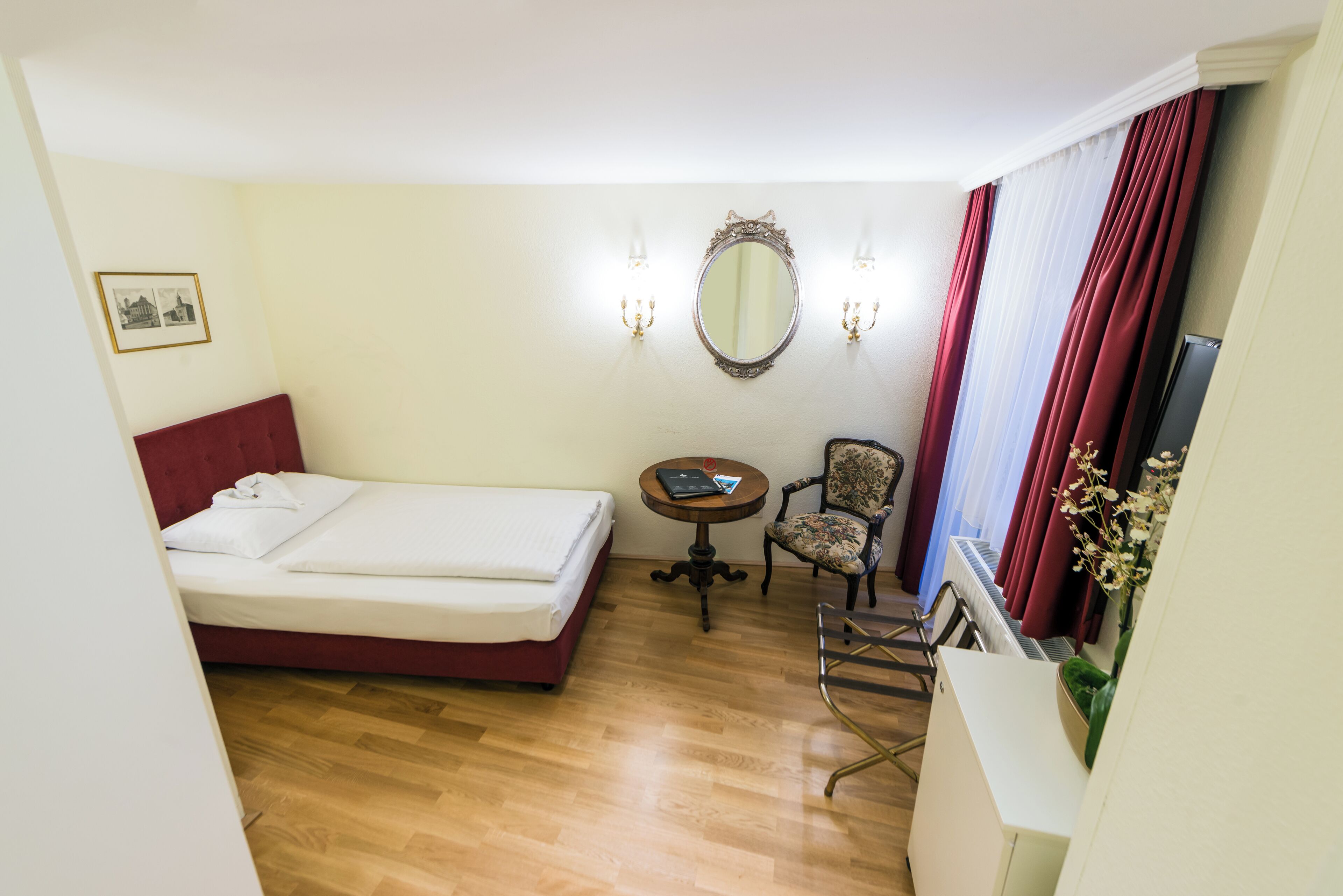 A hotel room with a bed, a small table, a chair, a mirror, and a radiator.