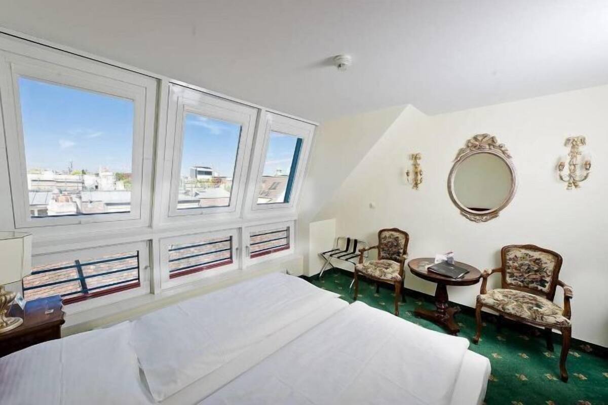 Hotellihuone, jossa on sänky, kaksi tuolia, pieni pöytä, peili ja suuret ikkunat, joista on näkymä rakennuksiin.