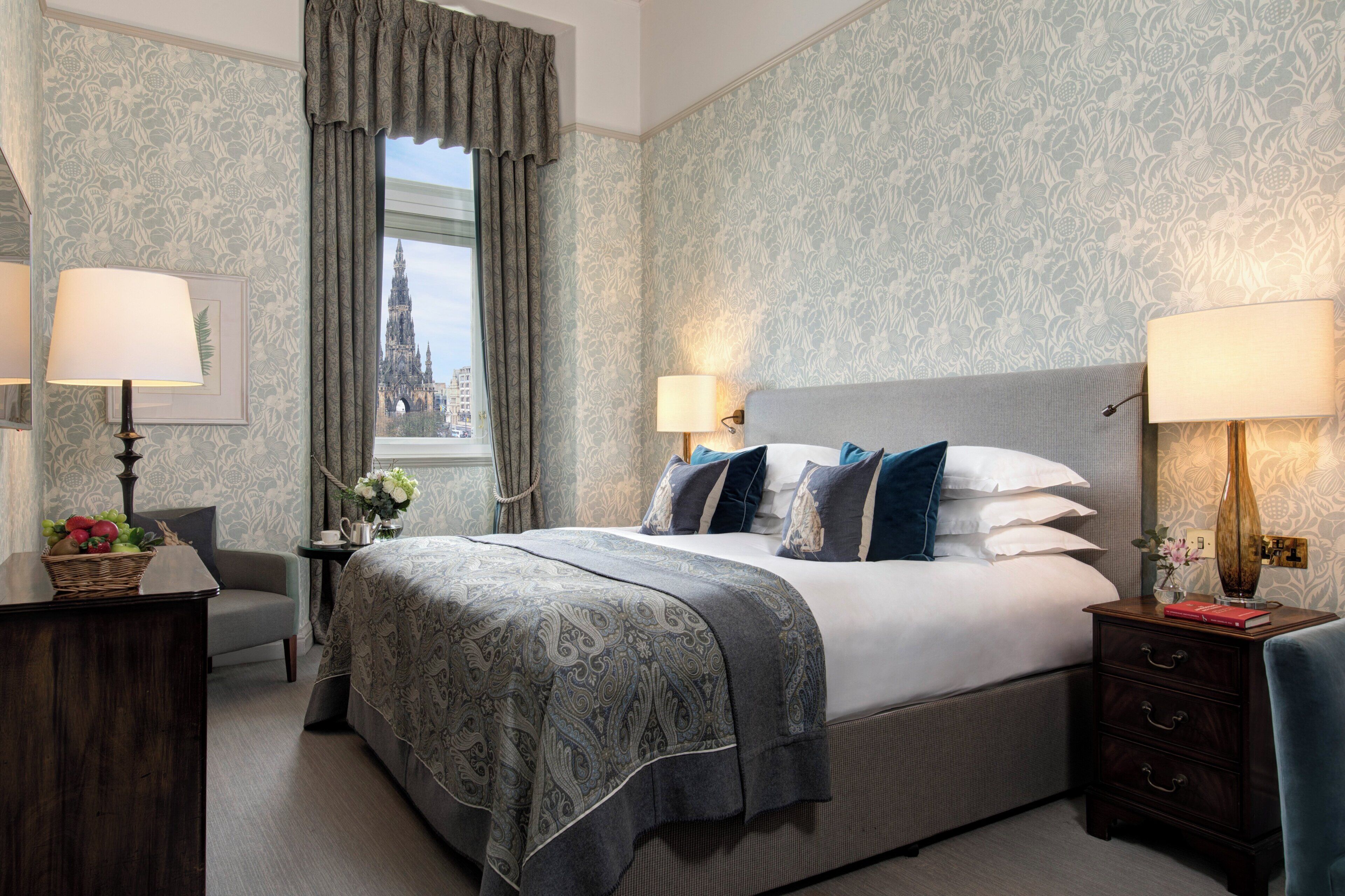 Quarto de hotel com uma cama grande, duas mesas de cabeceira, um criado-mudo, uma janela com vista para uma catedral e papel de parede estampado.