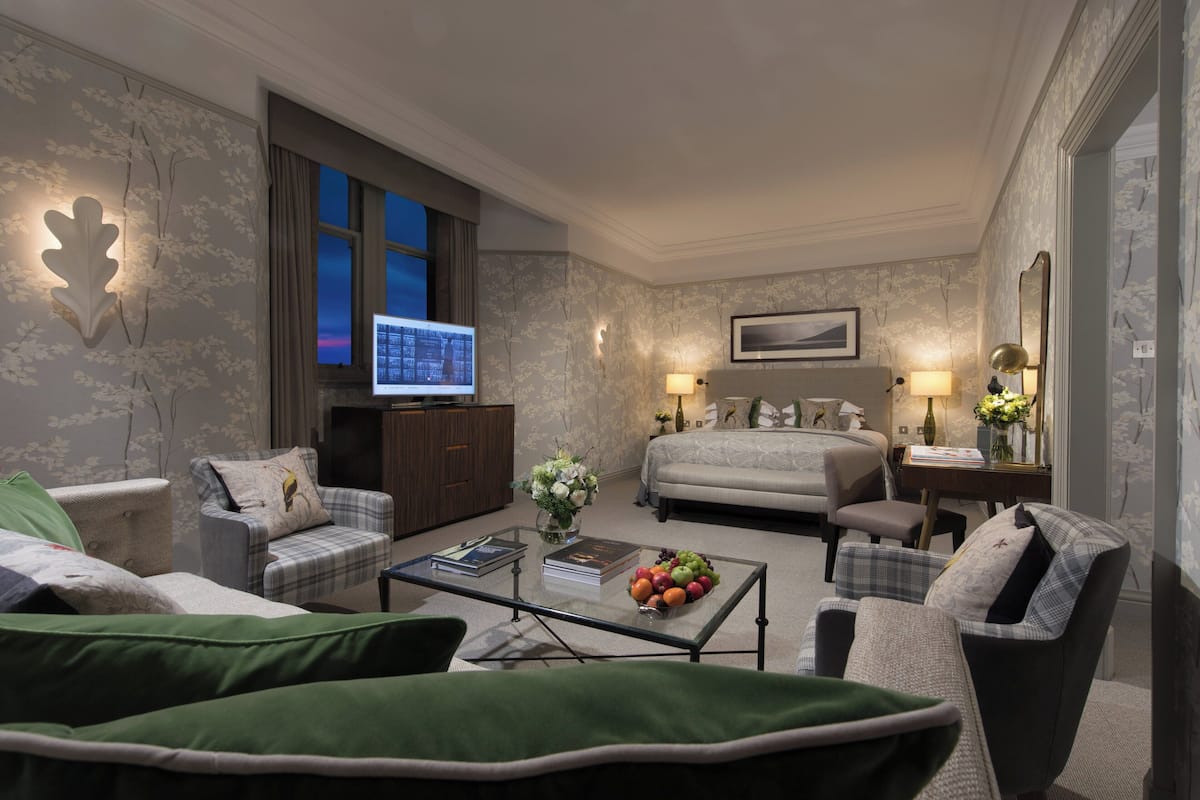 Camera d'albergo con un letto, due poltrone, un divano, un tavolino da caffè con libri e frutta, una televisione e vista sulla città.
