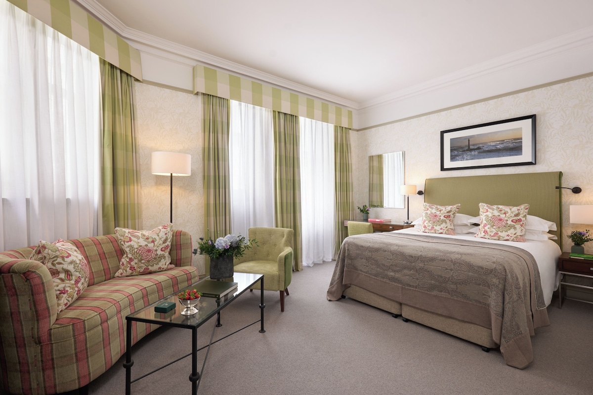 A hotel room with a bed, a sofa, a chair, a nightstand, a lamp, and a picture on the wall.