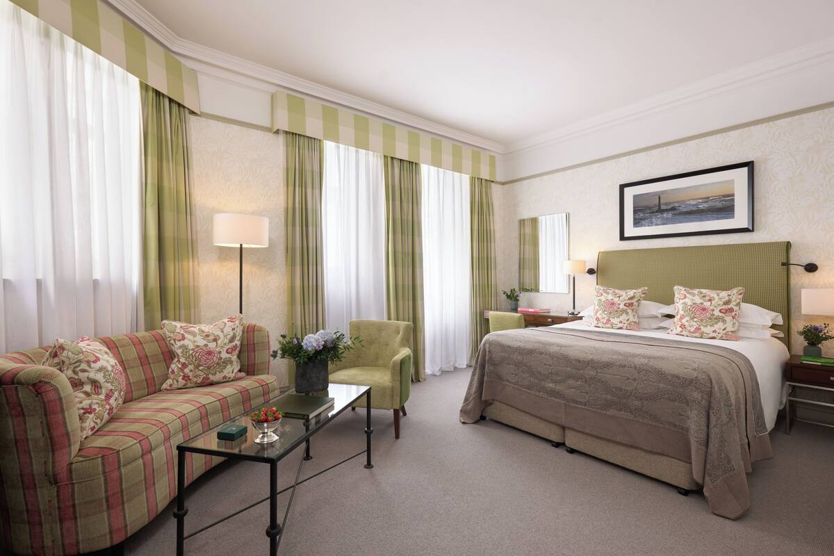 Una camera d'albergo con un letto, un divano, una poltrona, un comodino, una lampada e un quadro appeso al muro.