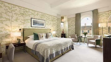 A hotel room with a large bed, a desk, a chair, a small table with a vase, and a view of a building through a window.