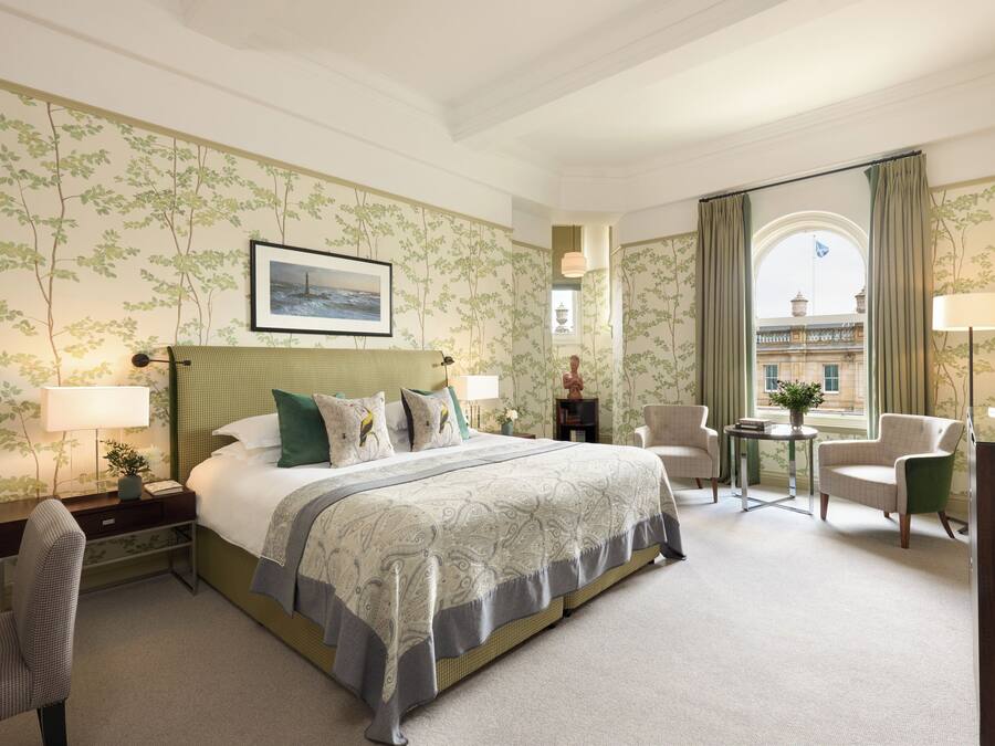 A hotel room with a large bed, a desk, a chair, a small table with a vase, and a view of a building through a window.
