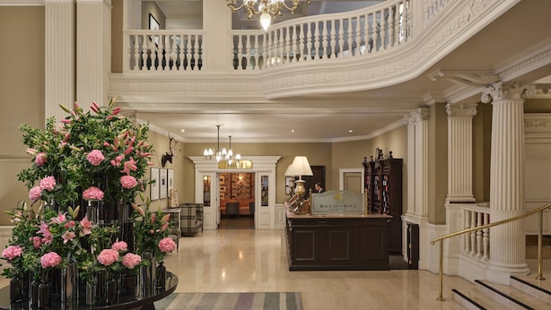 A grand lobby with a central floral arrangement, a reception desk, and a staircase leading to an upper level.