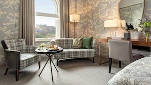 A hotel room with a plaid sofa, two armchairs, a round coffee table, a desk with a lamp, and a large mirror.