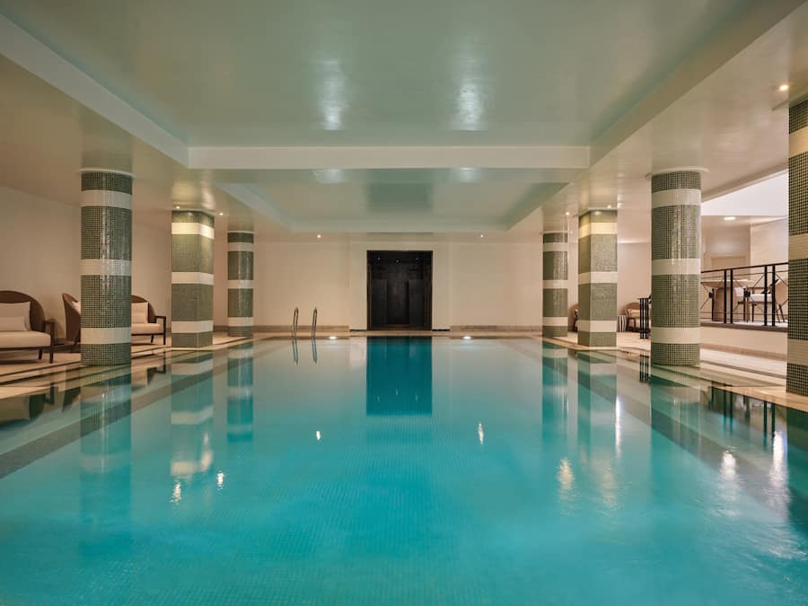 A large indoor swimming pool with clear blue water, surrounded by columns with mosaic patterns, and a reception area with seating.