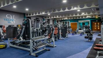 A gym with various exercise equipment, a wall with the text "VILLAGE Spa" and "HEALTH FITNESS BEAUTY", and a blue carpeted floor.