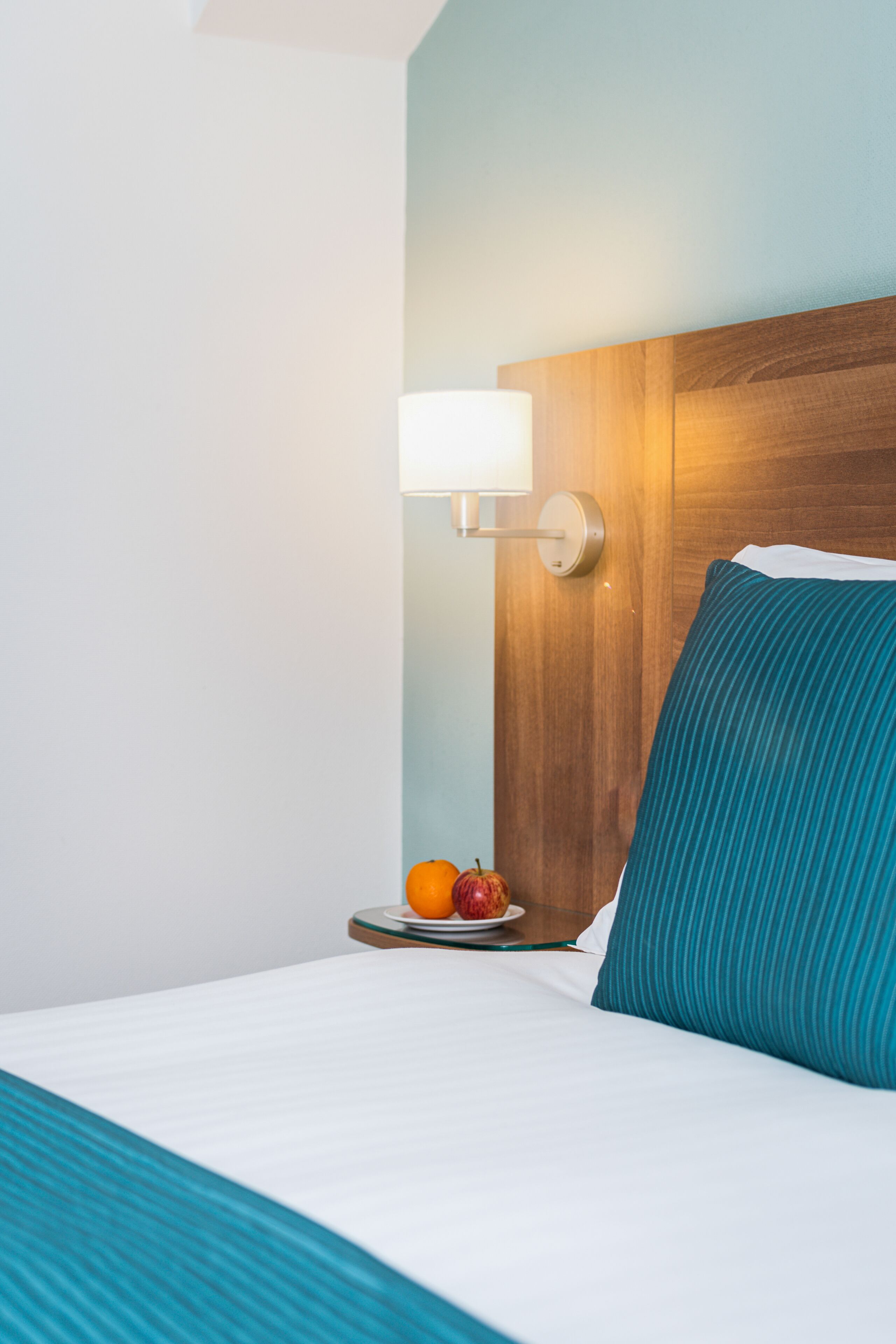 A neatly made bed with a teal pillow and a wooden headboard. A small table beside the bed holds an apple and a glass of water.