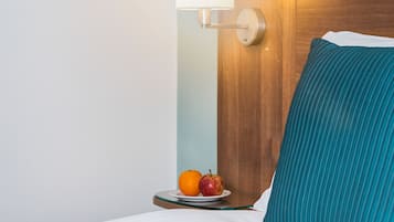 A neatly made bed with a teal pillow and a wooden headboard. A small table beside the bed holds an apple and a glass of water.