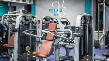 Un gimnasio con varias máquinas de ejercicio, una alfombra azul y una pared con el texto "VILLAGE Spa".