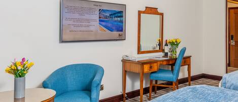 A hotel room with a bed, a desk with a chair, a small table, a vase with flowers, and a framed informational poster on the wall.