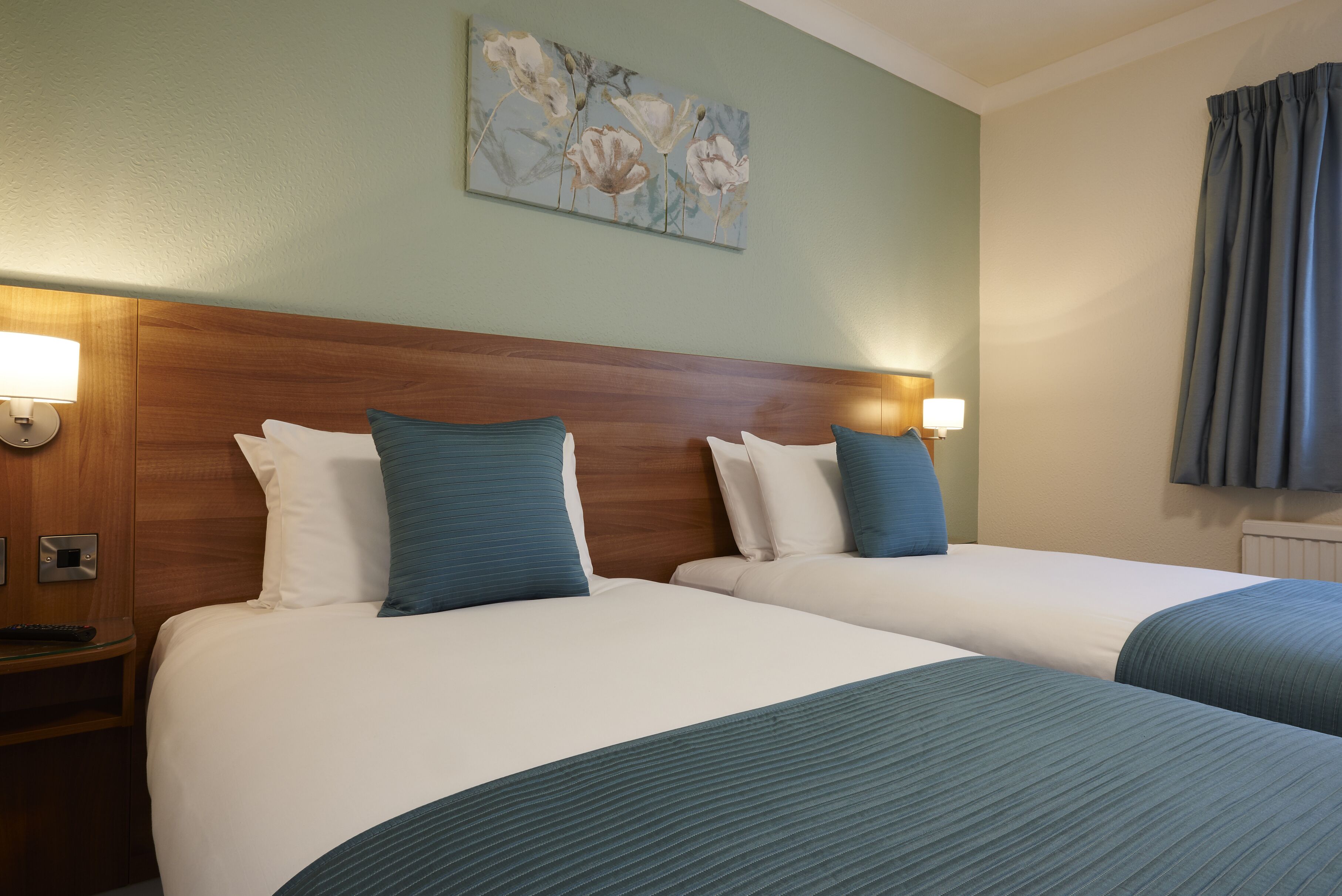 A hotel room with two beds, each with a wooden headboard, white bedding, and blue pillows. There is a wall-mounted painting above the beds and a nightstand with a lamp and a remote control.