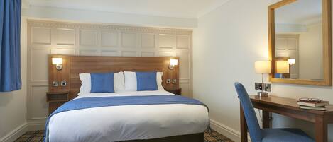 A hotel room with a large bed, a desk with a chair, a mirror, and a plaid-patterned floor.