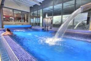 An indoor swimming pool with a water slide, clear blue water, and a tiled edge. - Last Drop Village Hotel and Spa (Bolton)