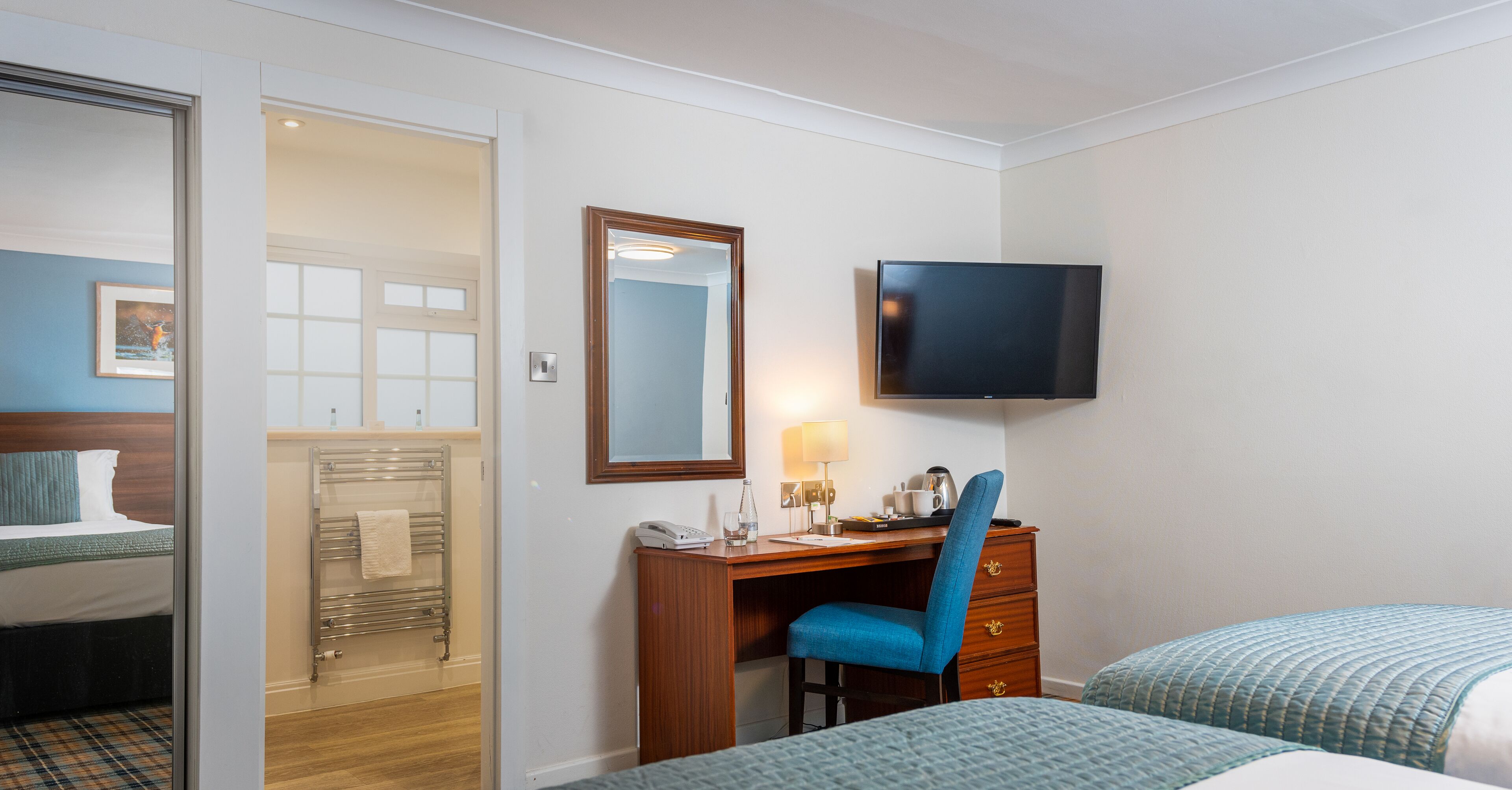 A hotel room with a bed, a desk with a chair, a TV mounted on the wall, and a bathroom visible through an open door.