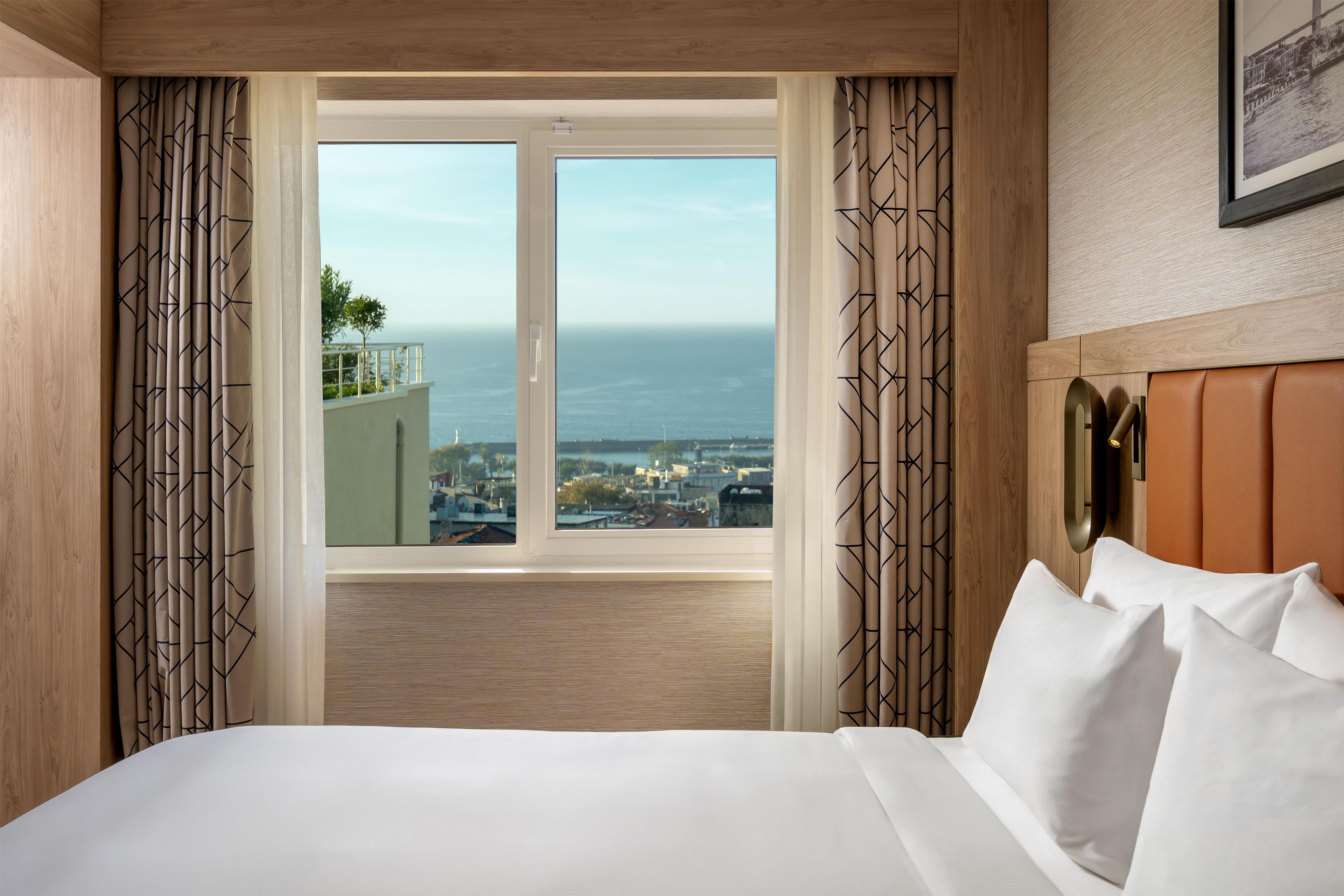 A hotel room with a large bed, a view of the city and sea, and patterned curtains.