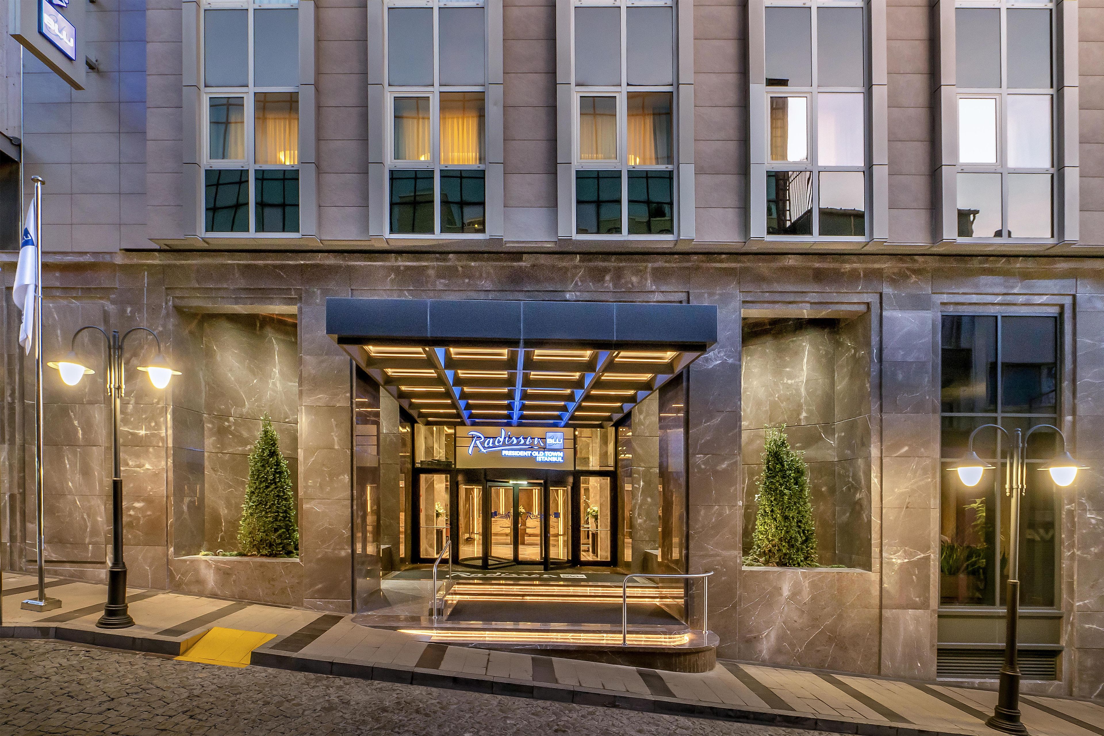 A hotel entrance with a glass door, a sign above the entrance, and two potted plants on either side.