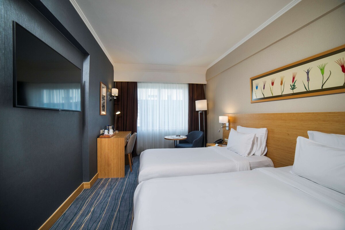 A hotel room with two beds, a desk, a chair, a TV, and a painting on the wall.