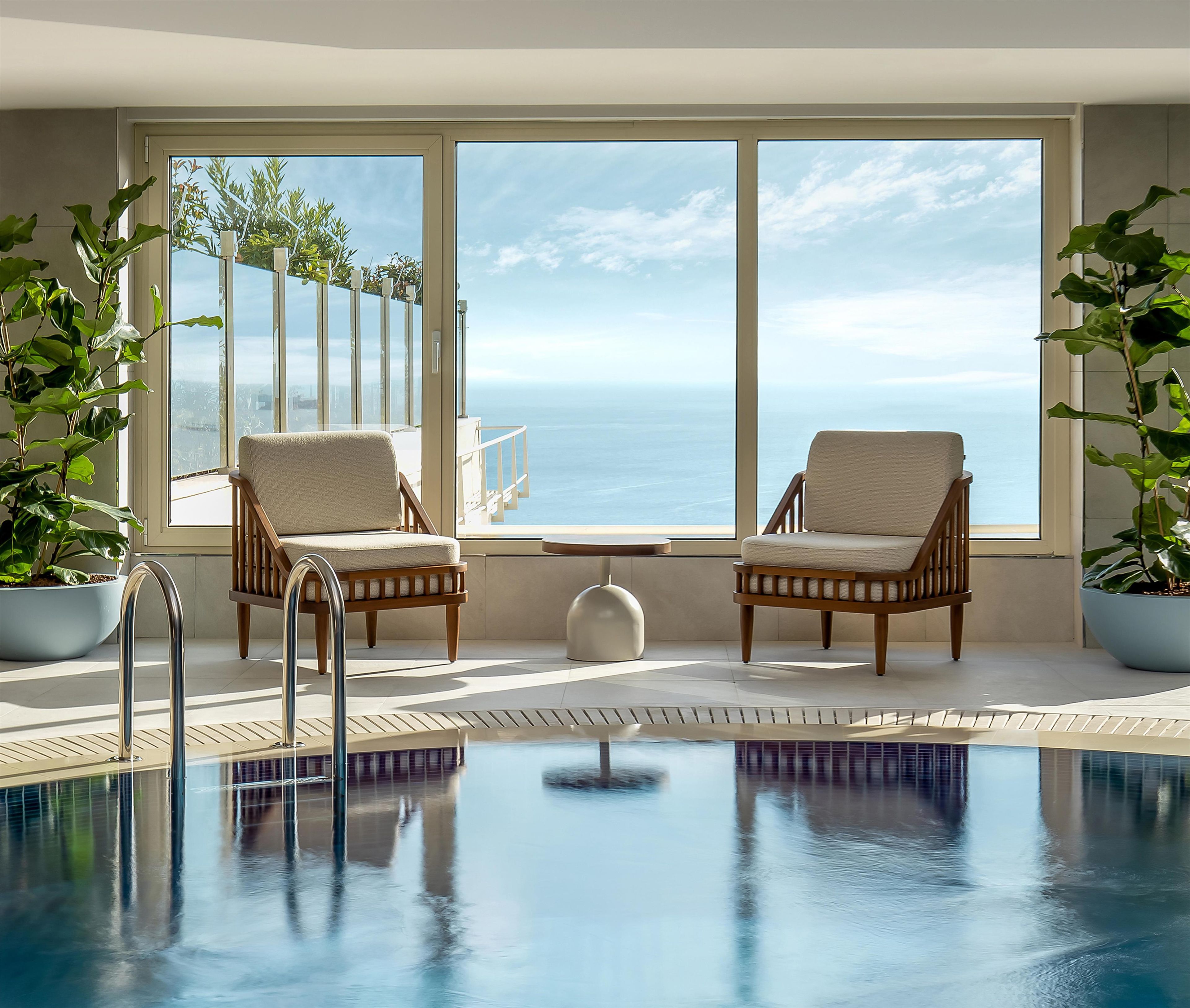 A pool area with two wicker chairs and a small table, large windows with a view of the sea, and potted plants.