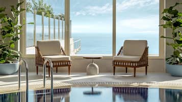 A pool area with two wicker chairs and a small table, large windows with a view of the sea, and potted plants.