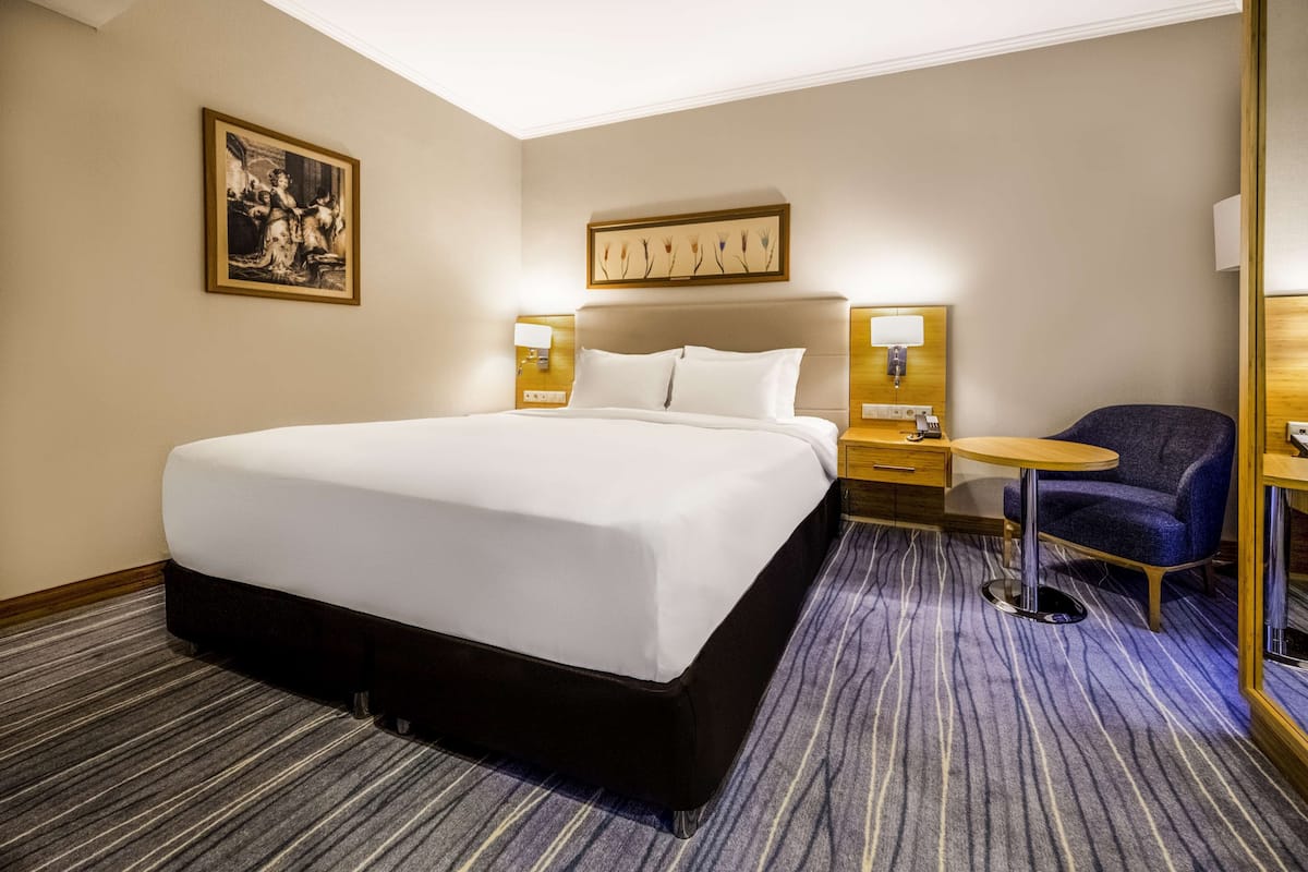 Habitación de hotel con una cama grande, dos mesitas de noche, una pequeña mesa redonda, una silla azul y dos lámparas de pared.