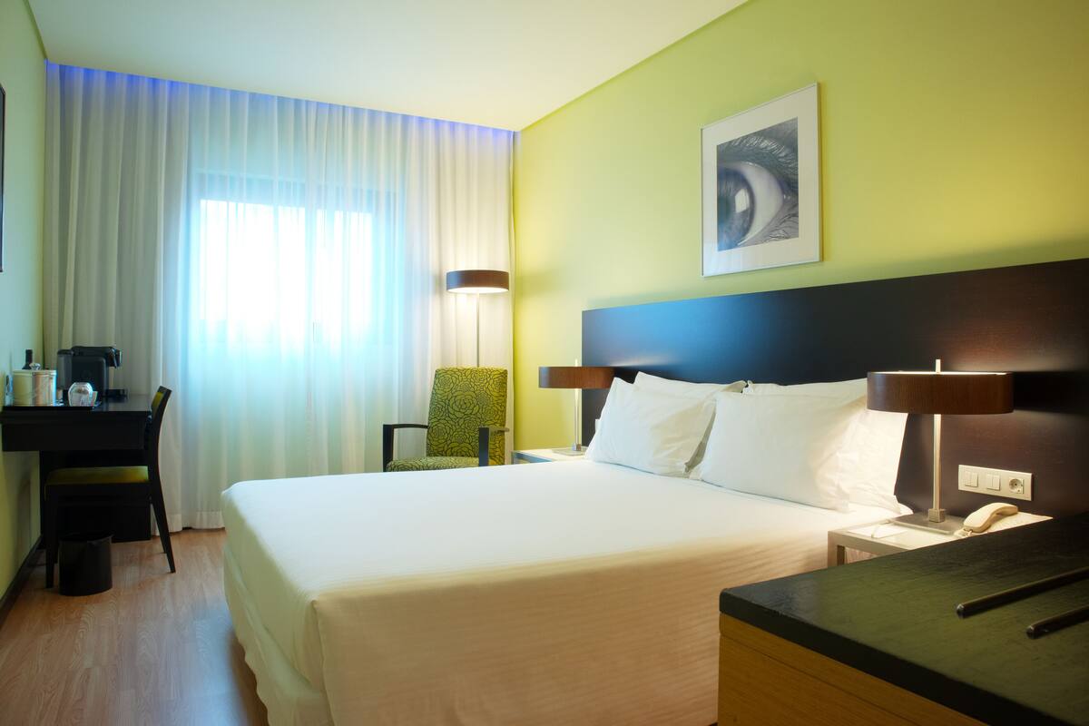 A hotel room with a bed, desk, chair, and a lamp.