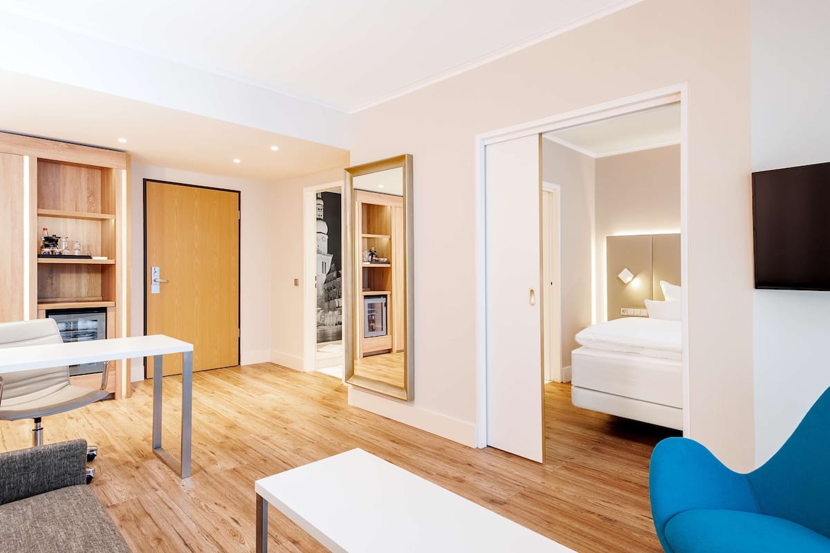 Una habitación de hotel moderna con piso de madera, una cama blanca, un sillón azul, un escritorio y estanterías empotradas de madera.